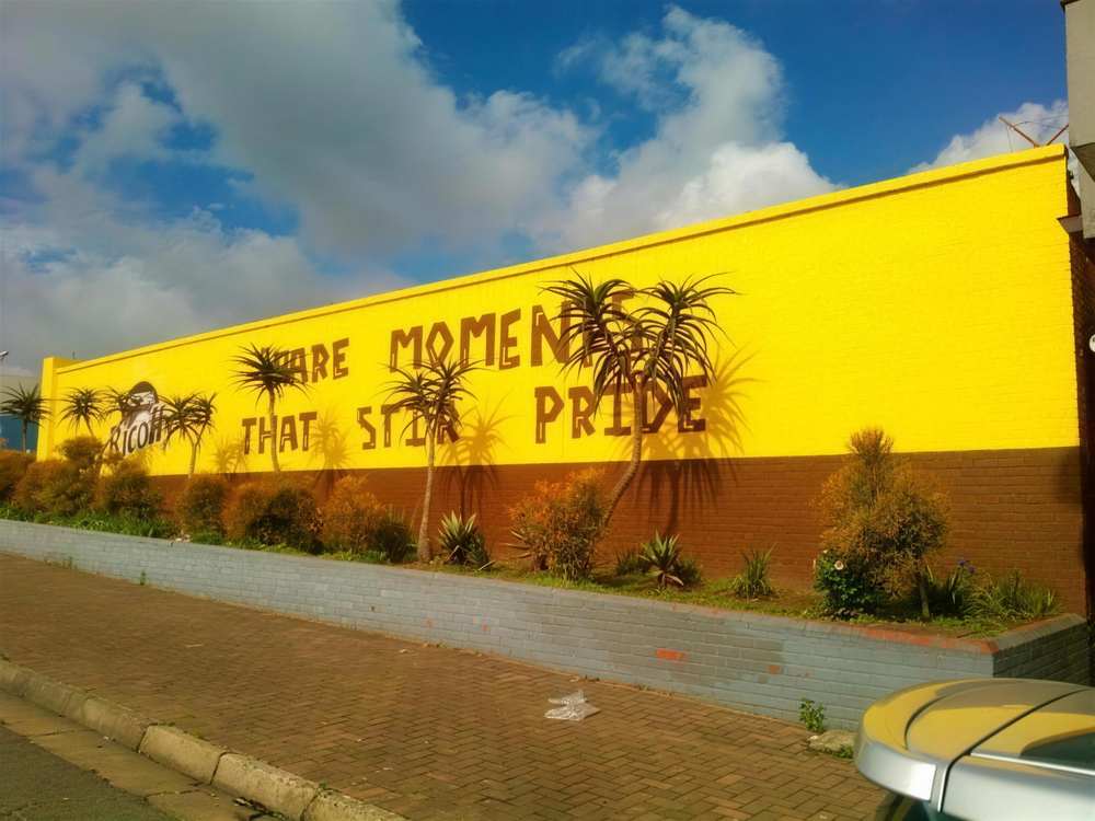 Bright yellow Ricoffy mural with painted aloes reading moments that stir pride